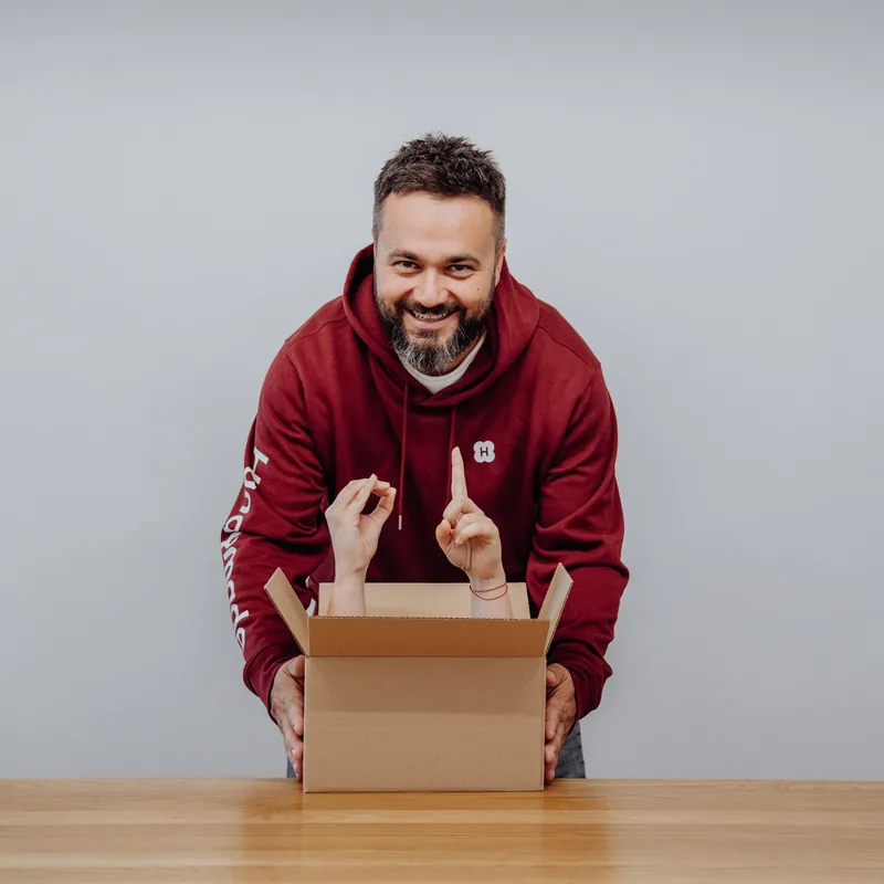 Z otevřené krabice vykukují dvě ruce, za ní se směje člověk z týmu v bordové mikině Handymade