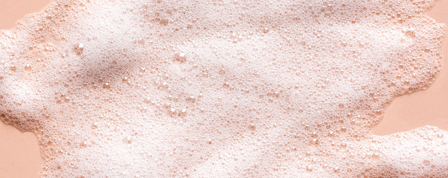 Biela pena s bublinkami vytvorená tenzidmi a surfaktantmi v kozmetickom produkte. Pena je na ružovom pozadí.